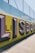 Large, bold graffiti-style letters spelling 'LISBOA' are painted on a concrete wall with a vibrant yellow background. The letters are shaded with purple and black, creating a three-dimensional effect. Above the wall, a metal fence can be seen in the bright daylight.