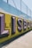 Large, bold graffiti-style letters spelling 'LISBOA' are painted on a concrete wall with a vibrant yellow background. The letters are shaded with purple and black, creating a three-dimensional effect. Above the wall, a metal fence can be seen in the bright daylight.