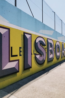 Large, bold graffiti-style letters spelling 'LISBOA' are painted on a concrete wall with a vibrant yellow background. The letters are shaded with purple and black, creating a three-dimensional effect. Above the wall, a metal fence can be seen in the bright daylight.