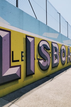 Large, bold graffiti-style letters spelling 'LISBOA' are painted on a concrete wall with a vibrant yellow background. The letters are shaded with purple and black, creating a three-dimensional effect. Above the wall, a metal fence can be seen in the bright daylight.