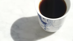 A white mug containing dark coffee is placed on a light marble surface. The mug has a blue artistic design, and its shadow is cast on the tabletop.