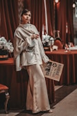 A woman is elegantly dressed in a silky, cream-colored outfit, holding a glass and leaning against a table draped in red fabric. She has a confident expression and is standing next to a vintage-style newspaper. The background includes rich red curtains and floral arrangements, enhancing a luxurious and sophisticated atmosphere.