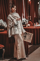 A woman is elegantly dressed in a silky, cream-colored outfit, holding a glass and leaning against a table draped in red fabric. She has a confident expression and is standing next to a vintage-style newspaper. The background includes rich red curtains and floral arrangements, enhancing a luxurious and sophisticated atmosphere.