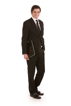 A person is wearing a formal black suit with a white shirt and a black tie, holding a closed laptop in one hand. The person is standing confidently and smiling, with their body slightly angled to the side against a white background.