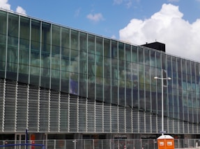 A modern glass building with a grid-like structure reflecting the sky and clouds. The lower part of the building has horizontal slats and there are entrances at the bottom. An orange portable toilet is visible in the foreground, along with construction barriers.