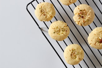 Round cookies with a crumbly texture are placed on a black metal cooling rack. Sprinkles of nuts or seeds are visible on the tops of the cookies, and the background is a plain, light gray surface.