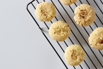 Round cookies with a crumbly texture are placed on a black metal cooling rack. Sprinkles of nuts or seeds are visible on the tops of the cookies, and the background is a plain, light gray surface.