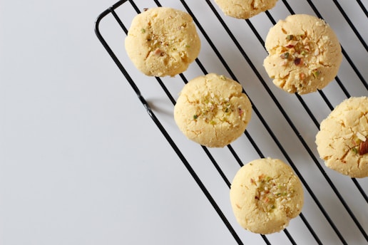Round cookies with a crumbly texture are placed on a black metal cooling rack. Sprinkles of nuts or seeds are visible on the tops of the cookies, and the background is a plain, light gray surface.