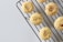 Round cookies with a crumbly texture are placed on a black metal cooling rack. Sprinkles of nuts or seeds are visible on the tops of the cookies, and the background is a plain, light gray surface.