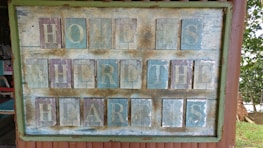 A rustic, weathered wooden sign with the words 'HOME IS WHERE THE HEART IS' painted in faded letters. The sign consists of individual wooden panels arranged in four rows, mounted on a wooden frame with visible signs of aging. The background shows natural surroundings, including trees and outdoor furniture.