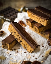 Thick chocolate-covered bars with a textured base featuring oats and possibly raisins are arranged on a piece of crumpled parchment paper. The background includes a blurred view of a bar of chocolate and scattered oats around the bars.