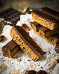 Thick chocolate-covered bars with a textured base featuring oats and possibly raisins are arranged on a piece of crumpled parchment paper. The background includes a blurred view of a bar of chocolate and scattered oats around the bars.