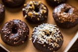 A tray of chocolate glazed donuts topped with various ingredients such as coconut shavings, chopped nuts, and chocolate bits. The donuts present a homemade and appetizing look, placed closely together on a baking sheet.