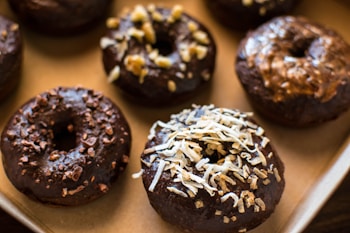 A tray of chocolate glazed donuts topped with various ingredients such as coconut shavings, chopped nuts, and chocolate bits. The donuts present a homemade and appetizing look, placed closely together on a baking sheet.