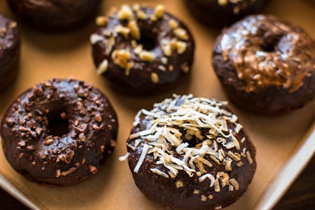 A tray of chocolate glazed donuts topped with various ingredients such as coconut shavings, chopped nuts, and chocolate bits. The donuts present a homemade and appetizing look, placed closely together on a baking sheet.