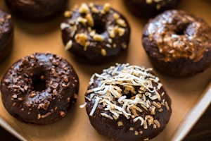 A tray of chocolate glazed donuts topped with various ingredients such as coconut shavings, chopped nuts, and chocolate bits. The donuts present a homemade and appetizing look, placed closely together on a baking sheet.