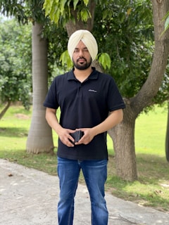 A man wearing a black polo shirt and blue jeans stands outdoors on a pathway. He is holding a smartphone and has a turban on his head. The background consists of green trees and grass, creating a serene, natural setting.