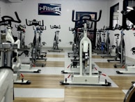 A room filled with stationary exercise bikes arranged neatly in rows. The space has a wooden and gray tiled floor, and a sign with the text 'i-Fitness' is displayed on the wall. The bikes are primarily white with black handlebars and seats.