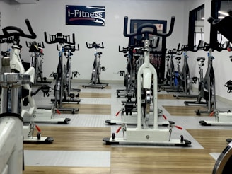 A room filled with stationary exercise bikes arranged neatly in rows. The space has a wooden and gray tiled floor, and a sign with the text 'i-Fitness' is displayed on the wall. The bikes are primarily white with black handlebars and seats.