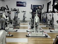 A room filled with stationary exercise bikes arranged neatly in rows. The space has a wooden and gray tiled floor, and a sign with the text 'i-Fitness' is displayed on the wall. The bikes are primarily white with black handlebars and seats.