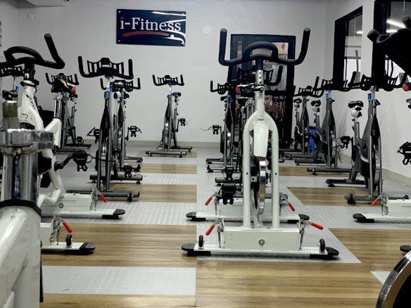A room filled with stationary exercise bikes arranged neatly in rows. The space has a wooden and gray tiled floor, and a sign with the text 'i-Fitness' is displayed on the wall. The bikes are primarily white with black handlebars and seats.