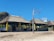 Traditional thatched-roof huts are situated on a sandy beach. The structures are supported by wooden pillars and have rustic fences made of bamboo or similar material surrounding them. A bare, white tree stands prominently in the foreground, and the sky is clear and blue. Lush greenery is visible behind the huts, indicating a tropical or coastal setting.