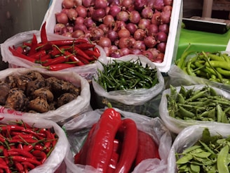 A market stall displays various fresh vegetables, including two types of red chili peppers, green chili peppers, green beans, mushrooms, large red peppers, and a container of red onions.