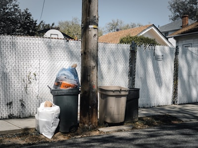 A suburban street scene with chain-link fencing surrounding a property. Three trash bins are positioned near a wooden utility pole, one overflowing with a blue bag on top. The atmosphere is quiet with residential houses in the background.