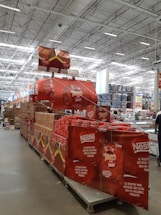 A large warehouse with high ceilings and bright lighting contains a neatly organized section of Nestlé chocolate products. Stacks of red and brown packaging are prominently displayed on shelves and pallets. Promotional signs and banners advertising the chocolate collection are visible.