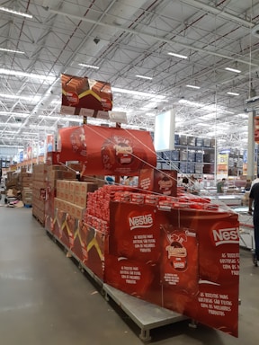 A large warehouse with high ceilings and bright lighting contains a neatly organized section of Nestlé chocolate products. Stacks of red and brown packaging are prominently displayed on shelves and pallets. Promotional signs and banners advertising the chocolate collection are visible.