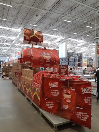 A large warehouse with high ceilings and bright lighting contains a neatly organized section of Nestlé chocolate products. Stacks of red and brown packaging are prominently displayed on shelves and pallets. Promotional signs and banners advertising the chocolate collection are visible.
