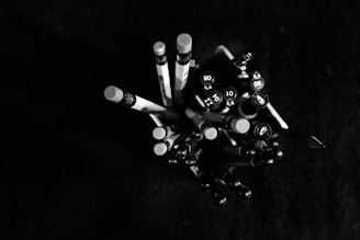 A cluster of pencils and pens is viewed from above, arranged in a container. The writing instruments vary in size and shape, with some showing erasers and others featuring pen tips. The image is primarily in black and white, highlighting the contrast between the objects and the dark background.