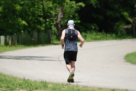 A person is running on a paved path surrounded by lush green trees. The individual is wearing a dark sleeveless shirt, black shorts, a light-colored cap, and running shoes. They appear to be jogging away, with the path curving ahead.
