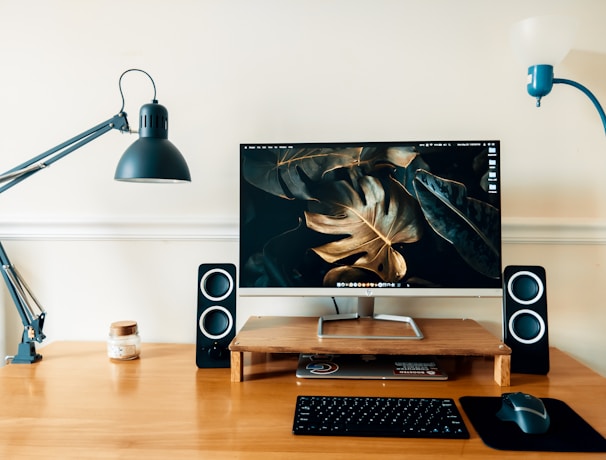 A neatly arranged wooden desk features a computer monitor on a wooden stand, displaying a wallpaper with dark, tropical leaves. On either side of the monitor are two small black speakers. To the left sits a grey adjustable desk lamp and a small jar candle. On the right, the desk has a mouse pad with a grey mouse on it. Above, there is a white and blue wall-mounted lamp.