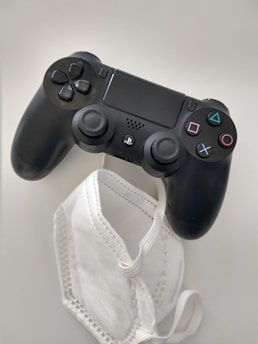 A black gaming controller is placed next to a white protective face mask on a light surface.