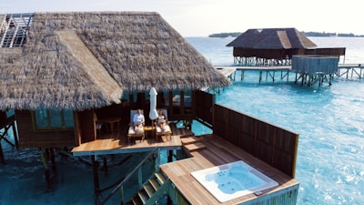 Two thatched-roof villas on stilts extend over clear turquoise waters. A wooden deck features a hot tub and two people relaxing on lounge chairs under an umbrella, enjoying the sun.