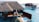Two thatched-roof villas on stilts extend over clear turquoise waters. A wooden deck features a hot tub and two people relaxing on lounge chairs under an umbrella, enjoying the sun.
