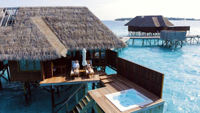 Two thatched-roof villas on stilts extend over clear turquoise waters. A wooden deck features a hot tub and two people relaxing on lounge chairs under an umbrella, enjoying the sun.
