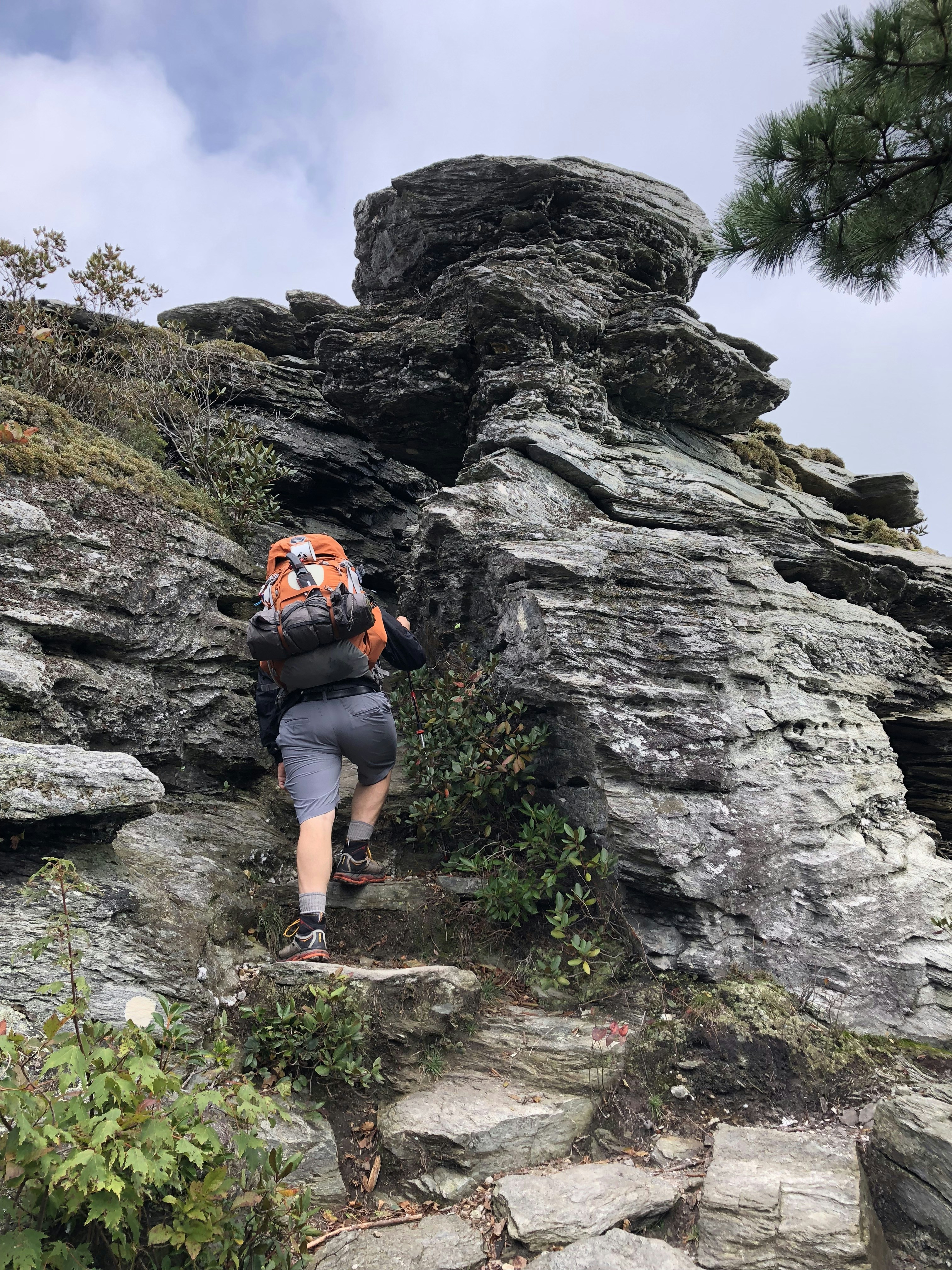 A person wearing a backpack climbs a steep rocky trail surrounded by rugged gray rock formations and sparse vegetation. The scene conveys a sense of adventure and exploration in a natural, slightly overcast setting.