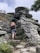A person wearing a backpack climbs a steep rocky trail surrounded by rugged gray rock formations and sparse vegetation. The scene conveys a sense of adventure and exploration in a natural, slightly overcast setting.