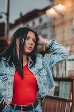 A woman with long dark hair wearing a denim jacket and a red top poses with one hand behind her head. She appears to be in an urban outdoor setting, possibly near a bookstore or newsstand, with blurred street elements and a light fixture in the background.