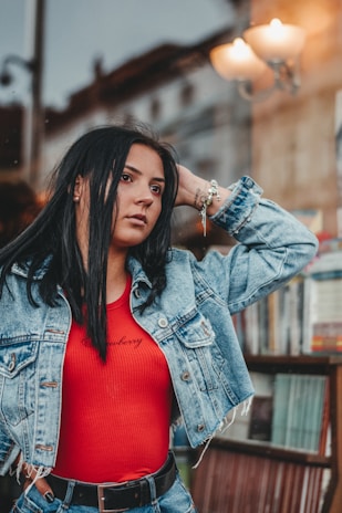 A woman with long dark hair wearing a denim jacket and a red top poses with one hand behind her head. She appears to be in an urban outdoor setting, possibly near a bookstore or newsstand, with blurred street elements and a light fixture in the background.