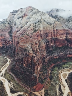 Aerial view of a rugged, multi-colored canyon with sheer rock cliffs and winding roads below. The canyon walls display layers of red, orange, and white rock, while greenery sparsely covers parts of the rugged landscape. River paths snake through the valley, and a cloudy sky looms overhead, adding depth and texture to the scene.