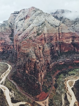 Aerial view of a rugged, multi-colored canyon with sheer rock cliffs and winding roads below. The canyon walls display layers of red, orange, and white rock, while greenery sparsely covers parts of the rugged landscape. River paths snake through the valley, and a cloudy sky looms overhead, adding depth and texture to the scene.