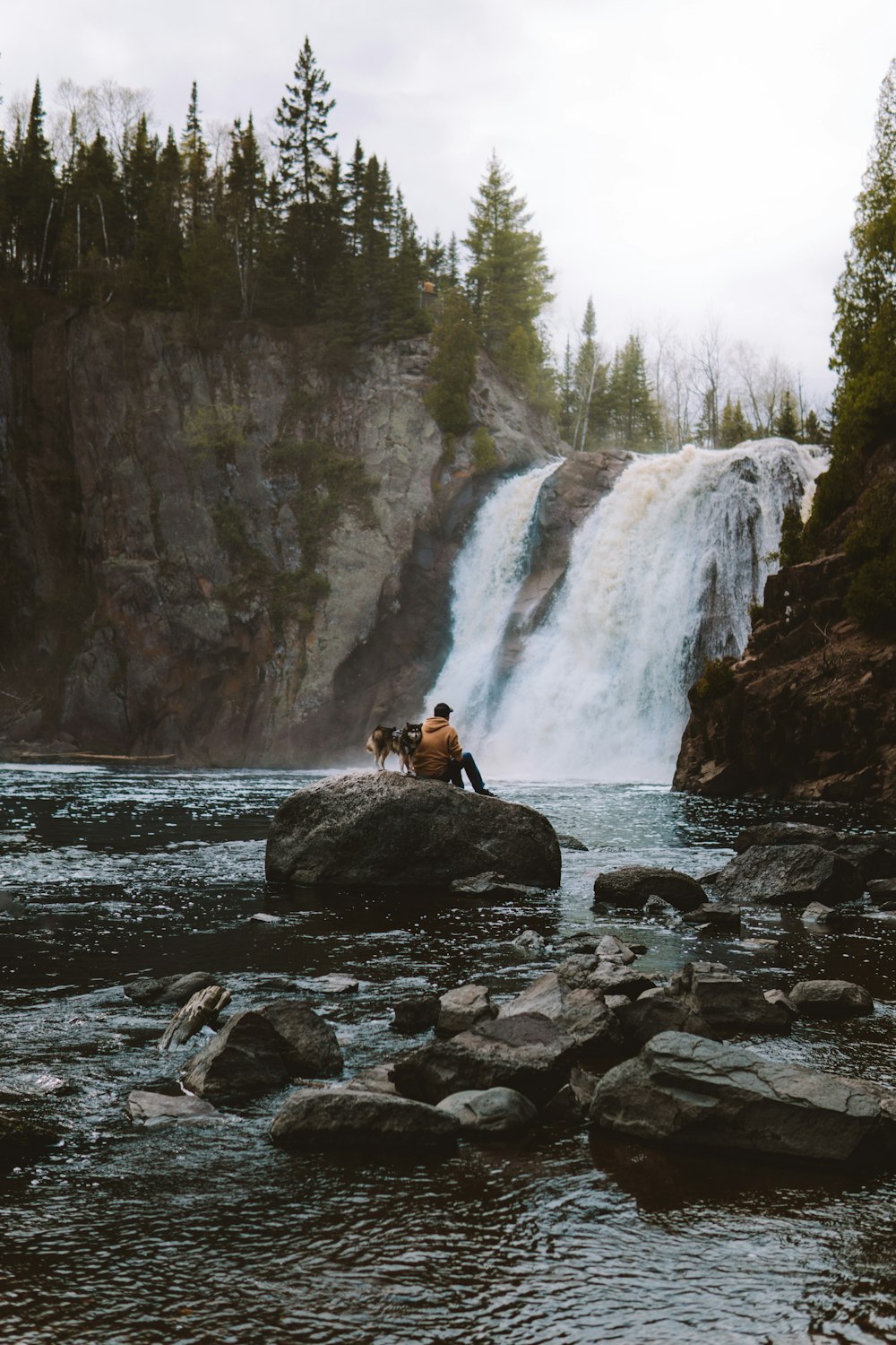 Frau im braunen Mantel sitzt tagsüber auf Felsen in der Nähe von Wasserfällen