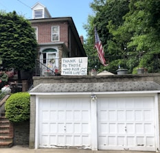 A brick house with a double garage is adorned with a handmade sign that reads 'Thank U to those who risk their life for us.' An American flag is displayed prominently beside the sign, and the house is surrounded by lush green trees and plants.