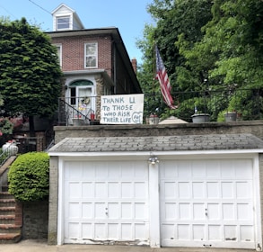 A brick house with a double garage is adorned with a handmade sign that reads 'Thank U to those who risk their life for us.' An American flag is displayed prominently beside the sign, and the house is surrounded by lush green trees and plants.