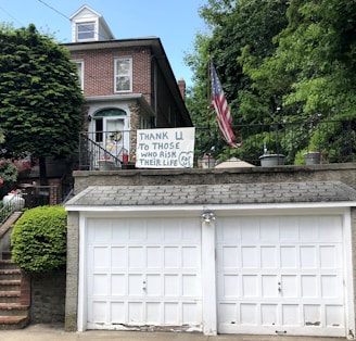A brick house with a double garage is adorned with a handmade sign that reads 'Thank U to those who risk their life for us.' An American flag is displayed prominently beside the sign, and the house is surrounded by lush green trees and plants.