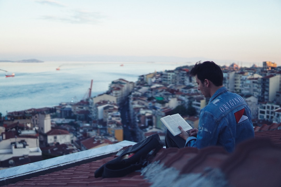 man in blue jacket reading book on top of building during daytime
