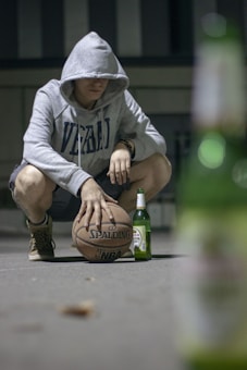 A person wearing a gray hoodie with the letters 'VERBAL' crouches on a concrete surface. Their hand rests on a basketball with the Spalding logo visible. A green glass bottle, likely a beer, is placed nearby. The background appears to be a dimly lit urban setting.